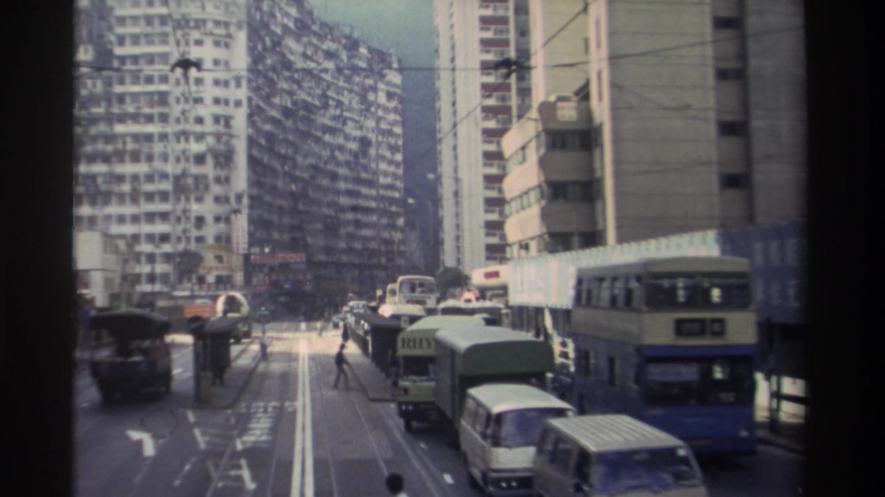 8mm footage from 1982 Hong Kong showing a wide multi-lane urban street with double-decker buses, vans, overhead tram wires, and dense residential towers flanking both sides.