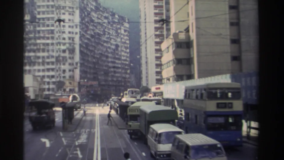 8mm footage from 1982 Hong Kong showing a wide multi-lane urban street with double-decker buses, vans, overhead tram wires, and dense residential towers flanking both sides.