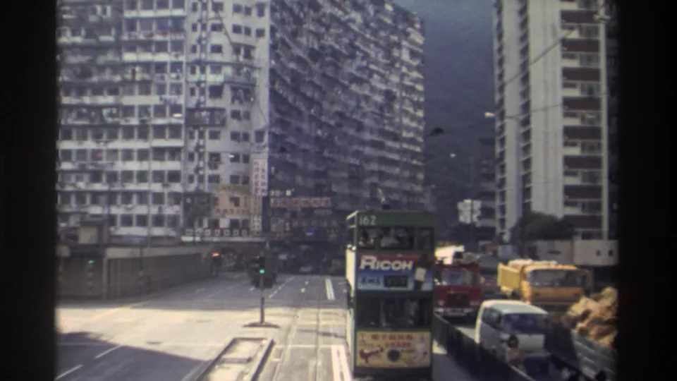 8mm footage from 1982 Hong Kong with double-decker tram 162 and its Ricoh panel prominent, green traffic signal showing, yellow construction equipment to the right, and dense tower blocks in background.