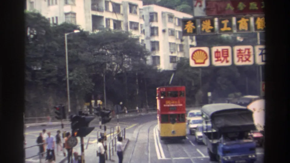 Hong Kong, c.1982: tram moving through busy intersection flanked by period vehicles; Shell signage and Chinese-character signs visible on buildings.