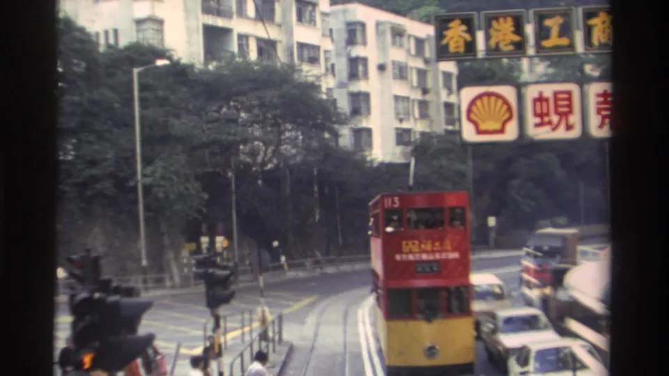 Hong Kong, c.1982: red and yellow double-decker tram dominates frame as it approaches; Shell logo and bilingual commercial signage visible overhead.