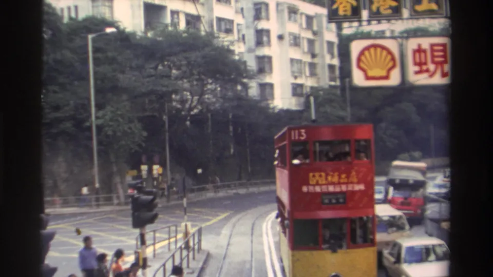 Hong Kong tram 113 navigating a curve with Shell hoarding and Chinese-language signs overhead, period sedans in traffic and pedestrians on the sidewalk, circa 1982, 8mm film.
