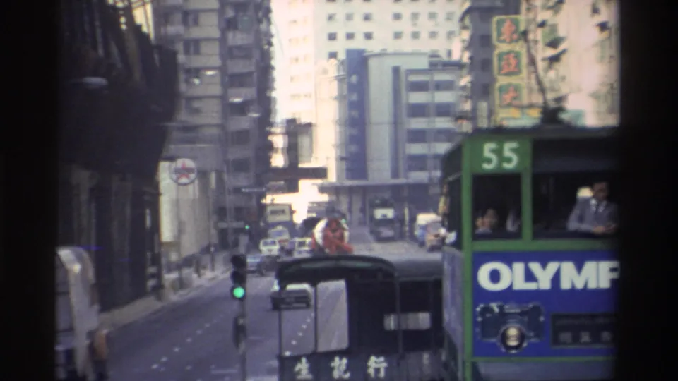 In-vehicle POV on a busy Hong Kong street, circa 1982; green bus 55 carrying a panel reading 'OLYMP', green traffic light, dense Chinese-language shopfronts and period sedans ahead.