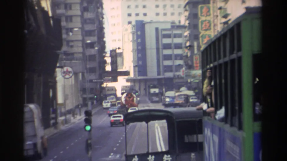 Handheld in-vehicle shot on a Hong Kong street, circa 1982; green bus with 'OLYMP' advertisement, green traffic light, Chinese-language shopfronts, driver visible in adjacent vehicle.