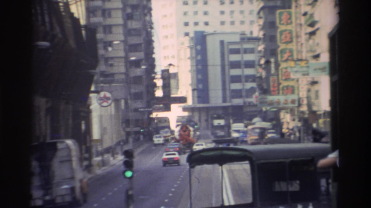 POV tram shot on a Hong Kong street in 1982, showing mixed traffic, illuminated Chinese-character signs on high-rise buildings, a green signal light, and the tram roof structure in frame.