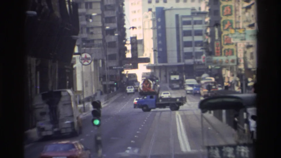 8mm POV from a Hong Kong tram in 1982 showing a blue flatbed truck amid mixed traffic, colorful Chinese-character signage on high-rise buildings, and a green traffic signal.