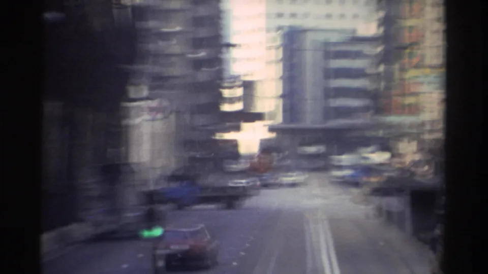 Motion-blurred 8mm frame from a tram in 1982 Hong Kong showing streaked high-rise facades and blurred traffic, capturing the speed and physical texture of the urban ride.
