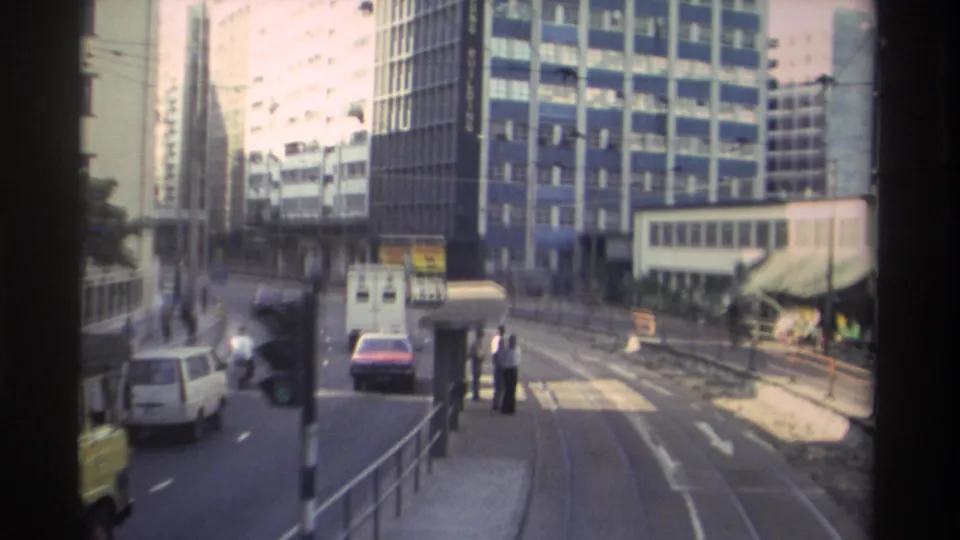 Late in the 1982 Hong Kong tram ride, the track curves near an open area with pedestrians visible on a roadside platform, a white van, and a prominent blue-grid-facade high-rise building.