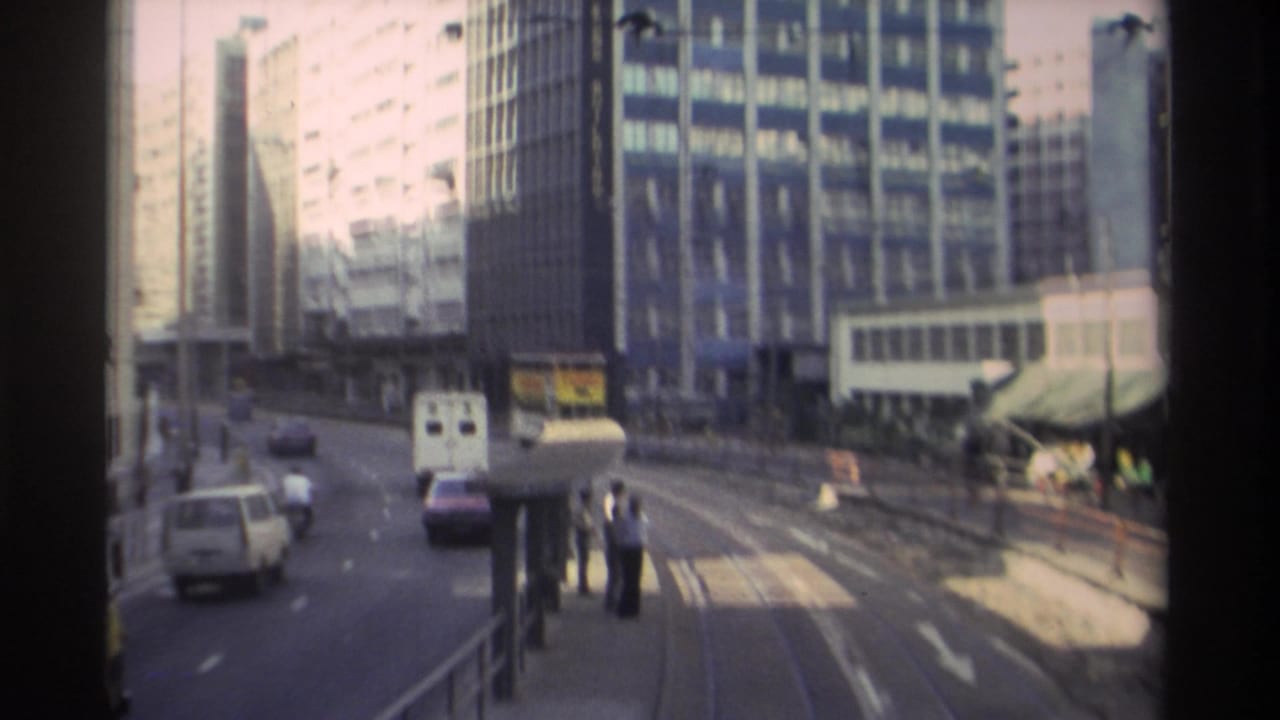 8mm footage of a wide Hong Kong boulevard circa 1982, showing tram tracks, high-rise buildings, a white van, and pedestrians near a roadside stop.