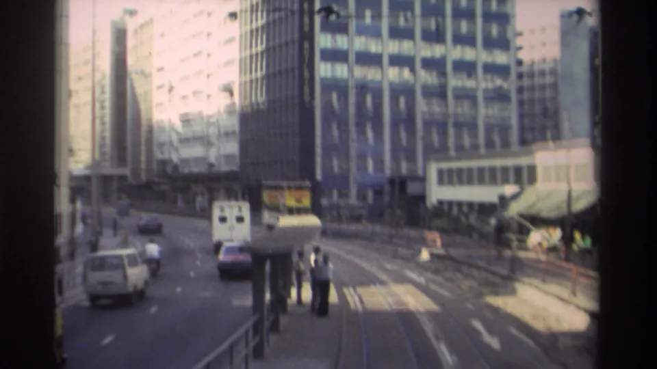 8mm footage of a wide Hong Kong boulevard circa 1982, showing tram tracks, high-rise buildings, a white van, and pedestrians near a roadside stop.