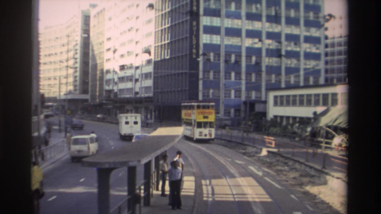 Hong Kong street with a yellow double-decker tram visible at right, two pedestrians near a traffic island, and tall buildings lining the boulevard.