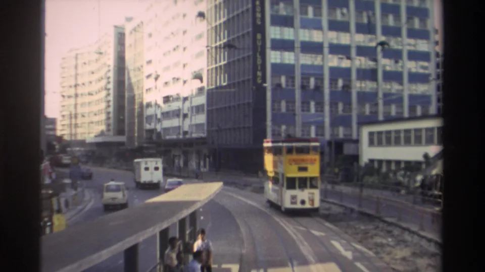 8mm archival frame from Hong Kong circa 1982; yellow double-decker tram with advertising panels travels curved tracks past modern office towers.