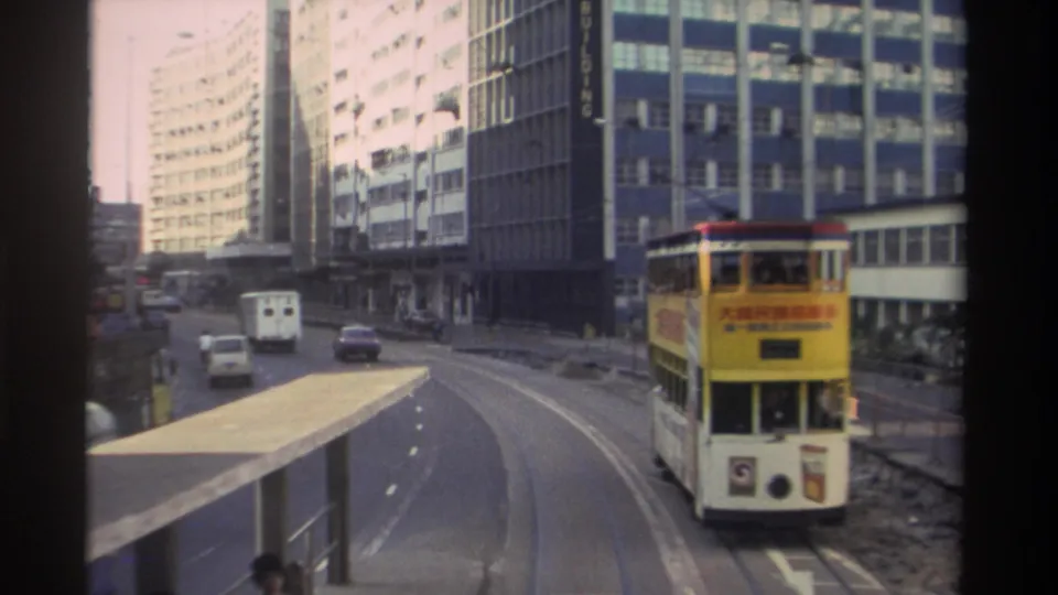 Close-medium 8mm frame from Hong Kong circa 1982; yellow double-decker tram with Chinese-text advertising panels curves through a wide boulevard lined with modern towers.