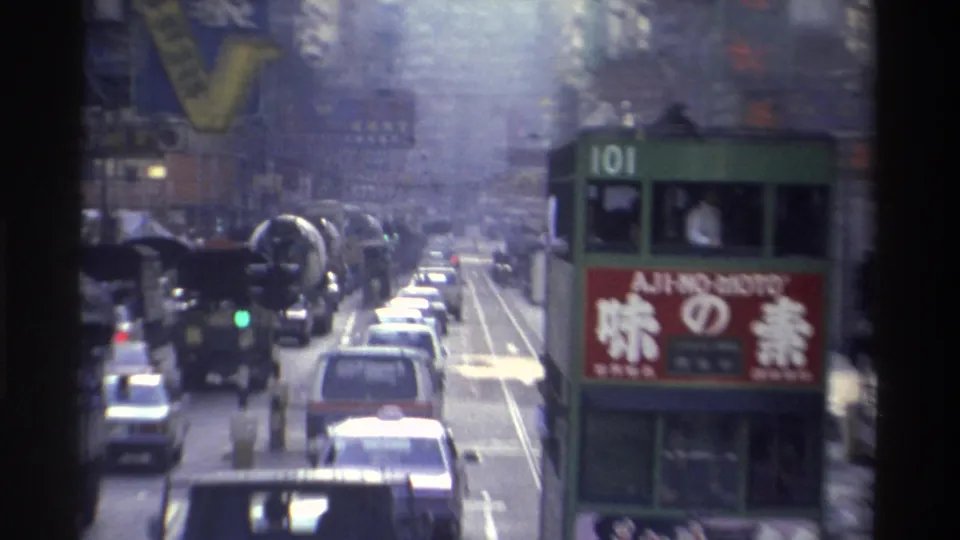1982 Hong Kong: tram route 101 with prominent AJI-NO-MOTO advertisement, surrounded by heavy traffic including trucks; high-rises lined with Chinese neon signage, Super 8 color shift visible.
