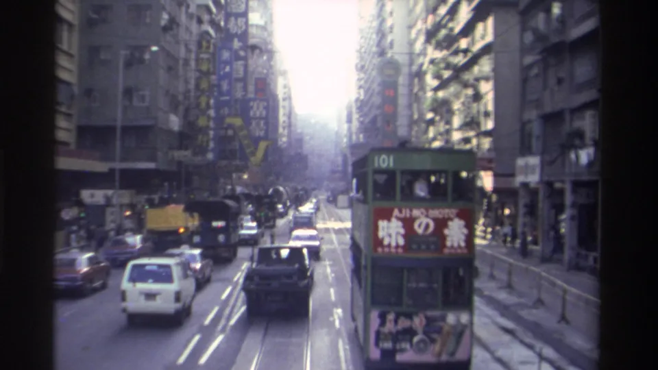 1982 Hong Kong street from elevated vantage, possibly tram upper deck; tram 101 ahead with Ajinomoto panel, yellow and white vehicles, Chinese neon signs, strong sun flare at horizon.