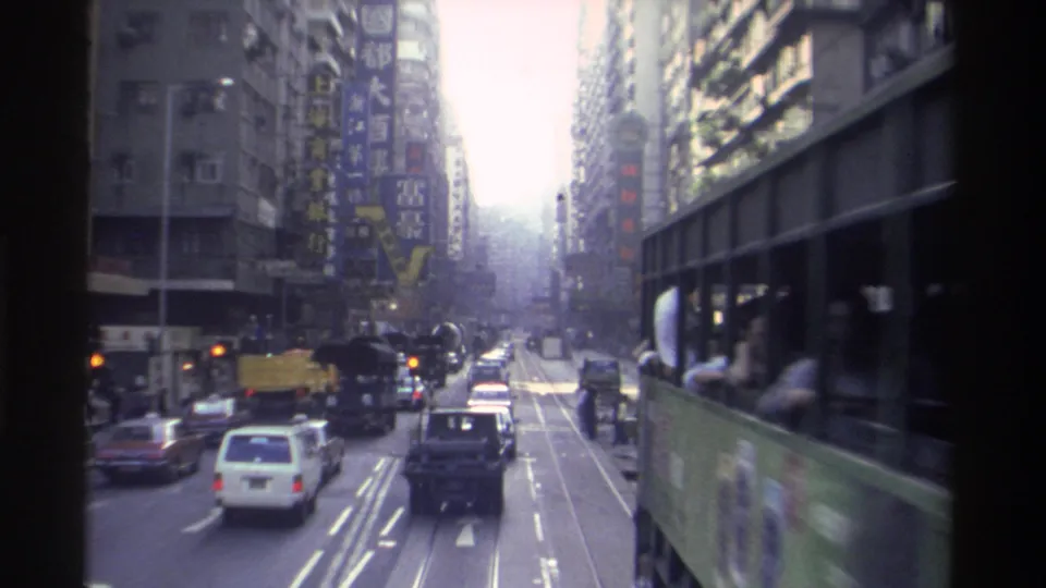 1982 Hong Kong: tram 101 passing at close range with side panel visible; white sedan in foreground, Chinese neon building signage, characteristic Super 8 blue-purple color shift.