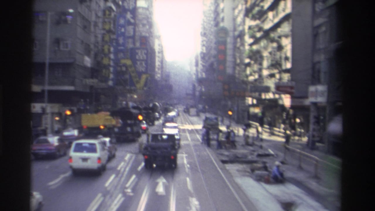 Hong Kong street viewed from a moving vehicle, showing tram tracks, cars, and high-rise buildings lining both sides.