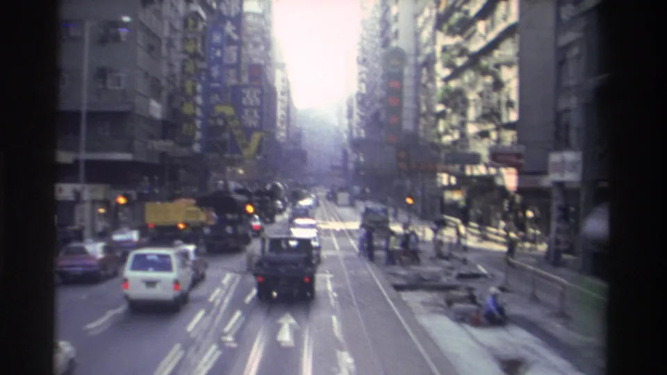 8mm footage of a busy Hong Kong commercial street circa 1982, with tram tracks, mixed traffic, high-rise buildings, and pedestrians visible.