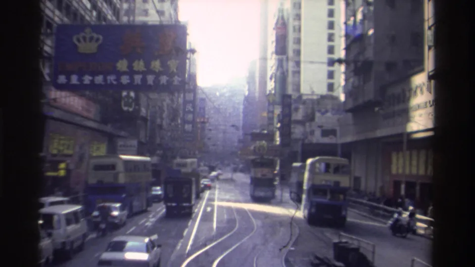 8mm street view of a Hong Kong commercial district circa 1982, with a double-decker tram, Chinese-language billboards, and a crown-logo commercial sign visible.