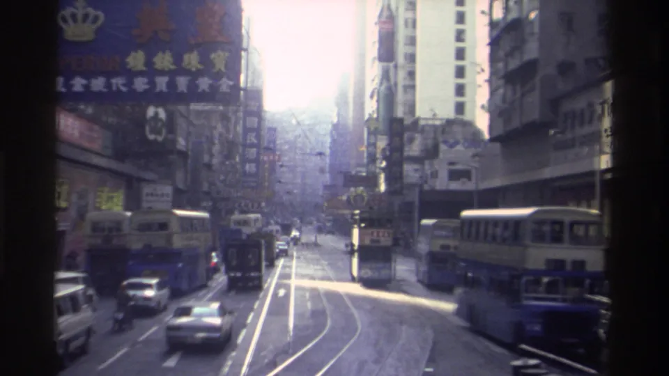 8mm footage of Hong Kong's commercial district circa 1982, with two double-decker trams, dense Chinese-language signage including a crown-logo sign, and tall buildings.