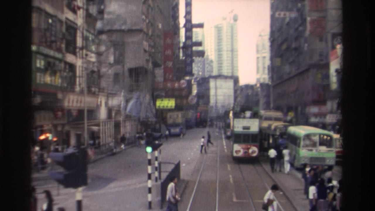 Busy Hong Kong street from an elevated angle showing double-decker trams, tram tracks, pedestrians crossing, and buildings with Chinese signage on both sides.