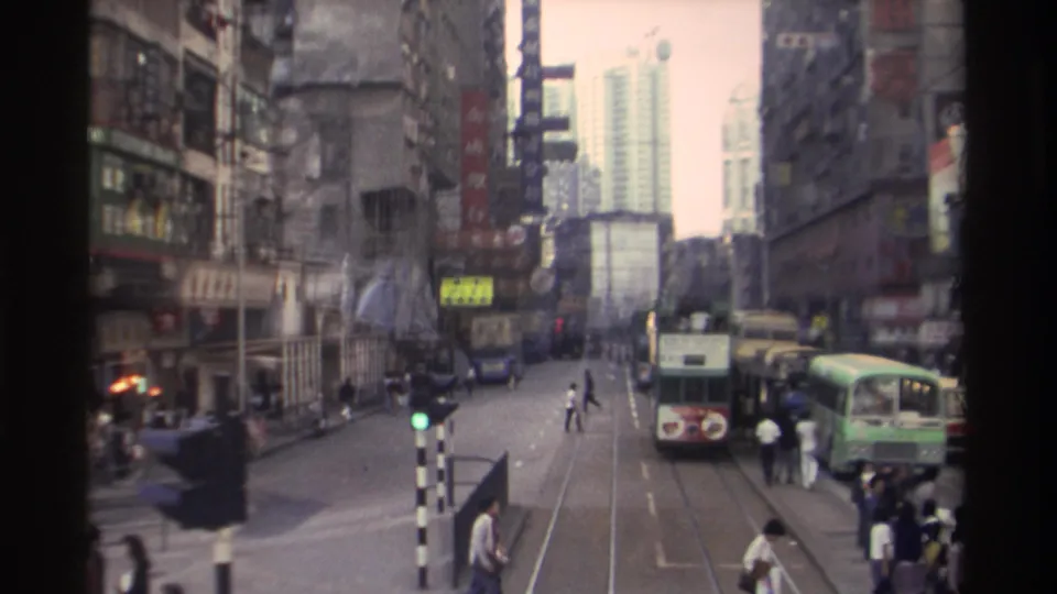 1982 Hong Kong street viewed from an elevated position: double-decker trams on embedded tracks, pedestrians crossing mid-road, and densely packed commercial buildings with Chinese and English signage.