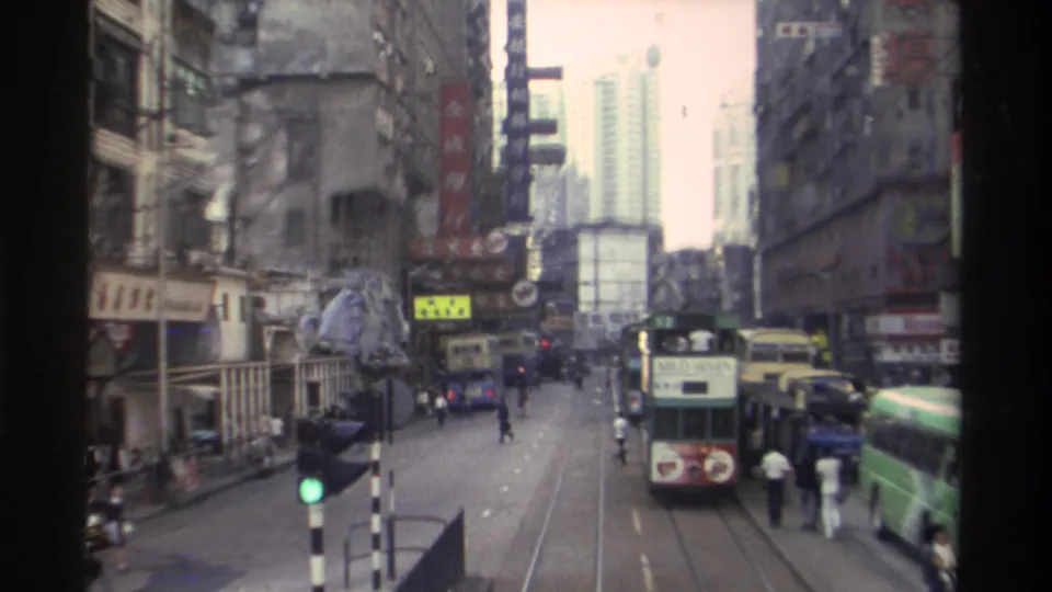 1982 Hong Kong: a green double-decker tram moves along embedded tram tracks past pedestrians, flanked by densely packed commercial buildings carrying Chinese and English signage.