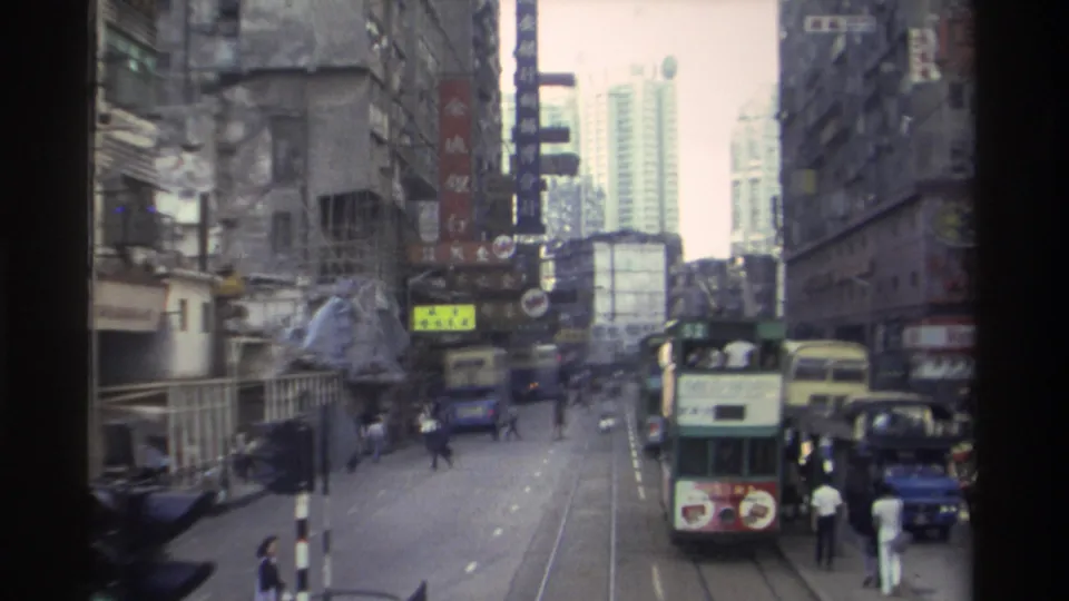 1982 Hong Kong: a green double-decker tram fills the foreground on embedded street tracks, surrounded by pedestrians and commercial buildings with Chinese-language signage, with a high-rise tower behind.