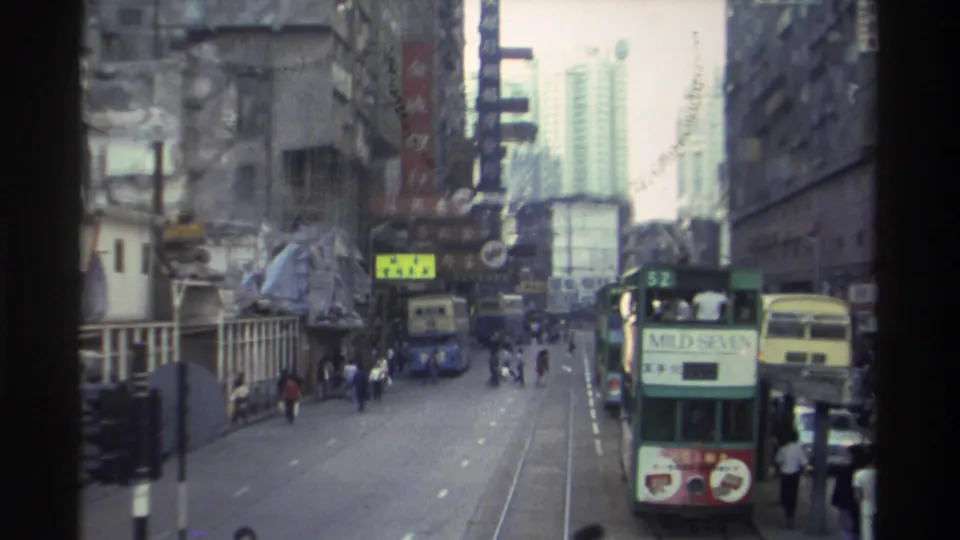 1982 Hong Kong: a double-decker tram displaying a Mild Seven cigarette advertisement and a route number passes in close view on a busy tram-track street, with pedestrians and Chinese-signage buildings behind.
