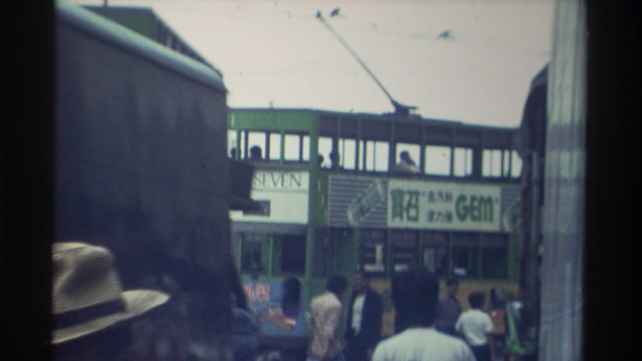 Close street view showing a green double-decker tram alongside shops; a person in a brimmed hat stands in foreground, crowd behind.