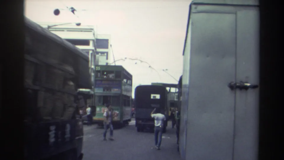 1982 Hong Kong urban street — white delivery truck partially fills foreground, green double-decker trams visible behind, pedestrian in white T-shirt.