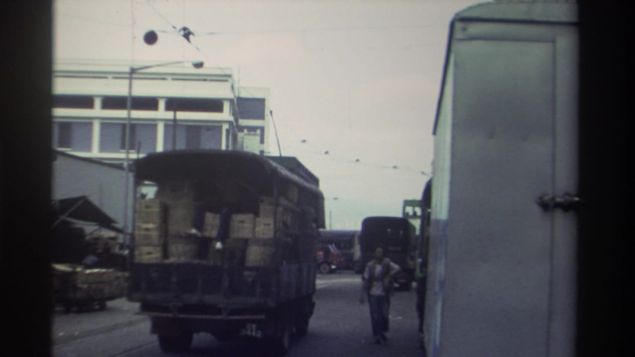Super 8 footage circa 1982 shows a crate-laden truck moving through a commercial Hong Kong street; a pedestrian walks alongside amid overhead wires and a concrete building.