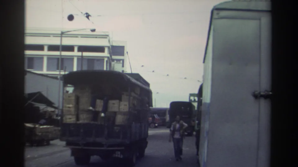 Super 8 footage circa 1982 shows a crate-laden truck moving through a commercial Hong Kong street; a pedestrian walks alongside amid overhead wires and a concrete building.