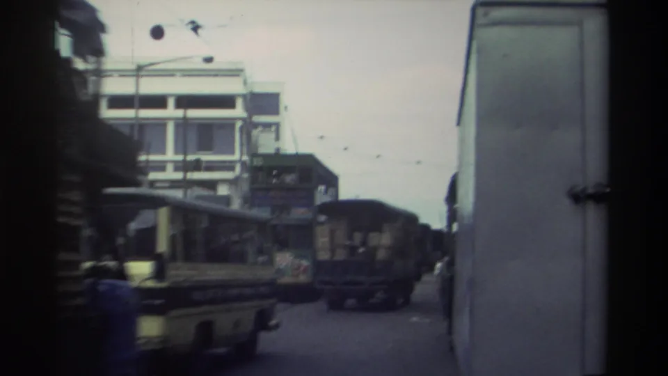 Hong Kong street circa 1982; a cream-colored bus and crate-loaded cargo trucks fill the frame, shot from inside an adjacent vehicle with characteristic Super 8 color fading.