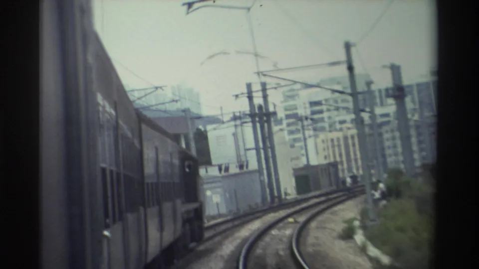 Train-level POV circa 1982 Hong Kong shows carriages stretching into the distance on an electrified rail line, with high-rise buildings under a hazy overcast sky.