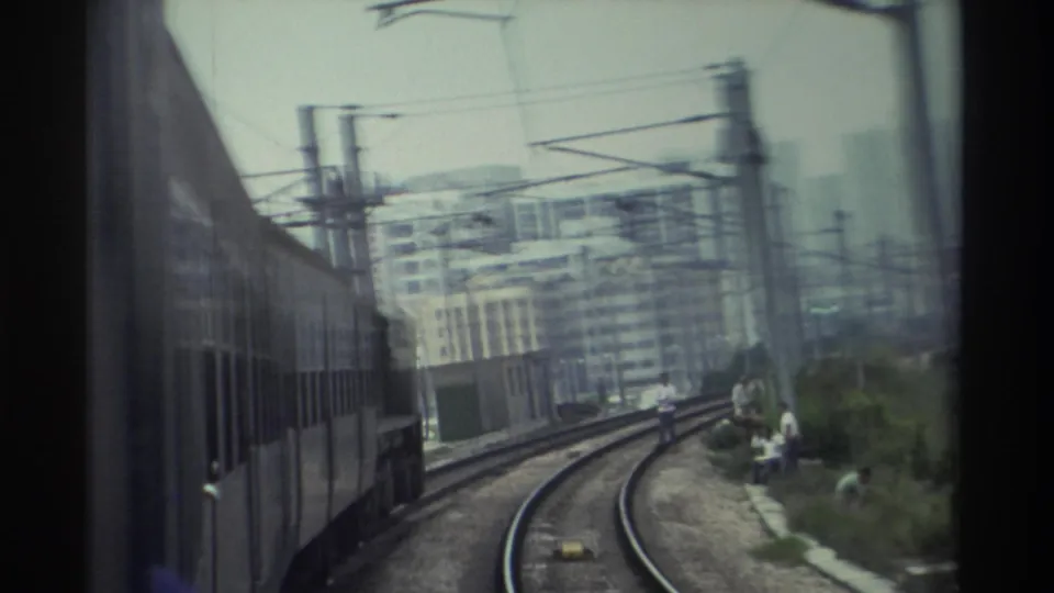 View from a moving train in Hong Kong, c. 1982, showing electrified rail infrastructure, residential towers, and several trackside figures.