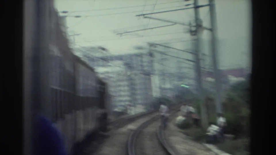 Motion-blurred frame from a moving train in Hong Kong, c. 1982, showing catenary infrastructure and figures alongside the curved track.