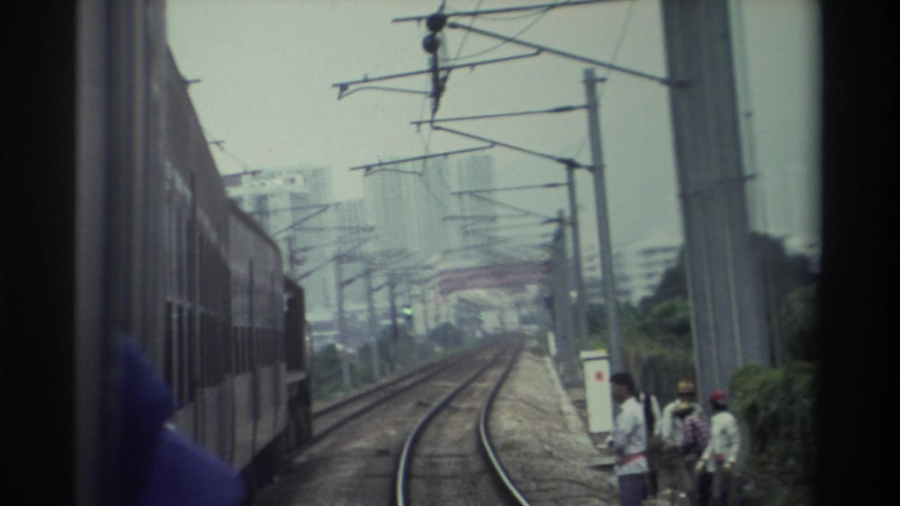 Super 8 POV from a moving train on what appears to be Hong Kong's electrified railway, 1982 — figures stand trackside, housing towers visible through haze.