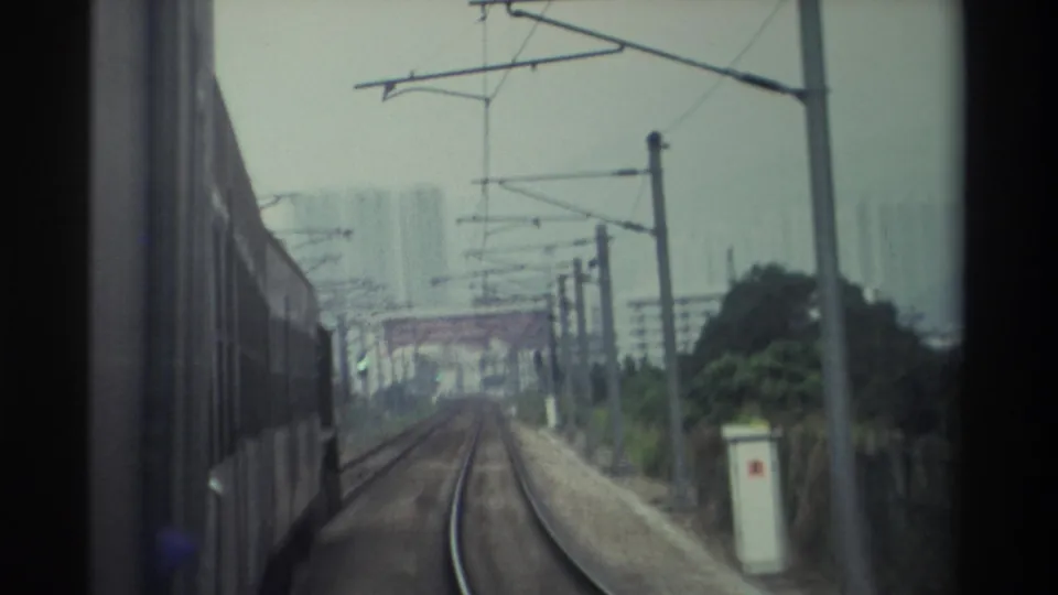 Super 8 train POV on Hong Kong railway, 1982 — overhead catenary infrastructure and distant high-rise residential blocks through overcast haze.
