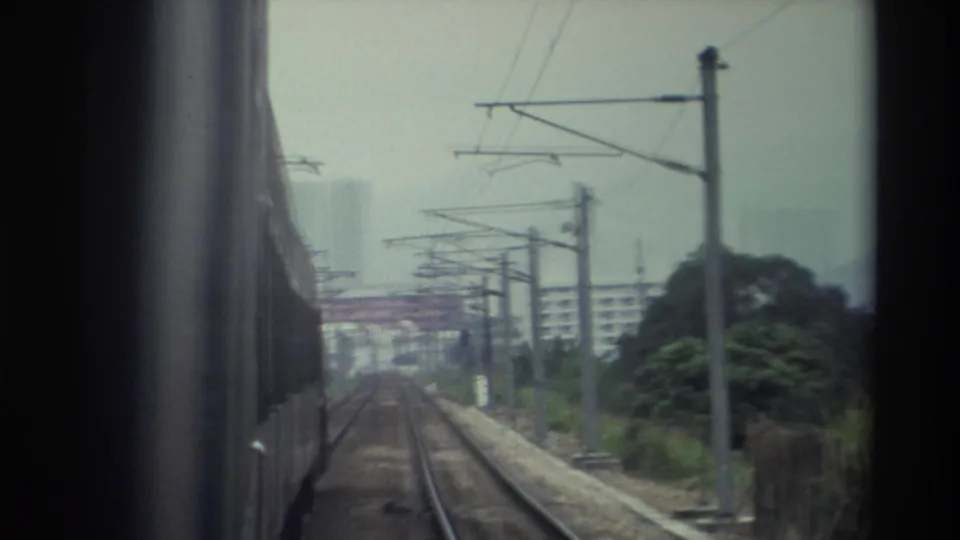 8mm POV from a moving train in what appears to be Hong Kong, 1982 — catenary and transmission pylons frame a track corridor with trees and hazy residential towers.