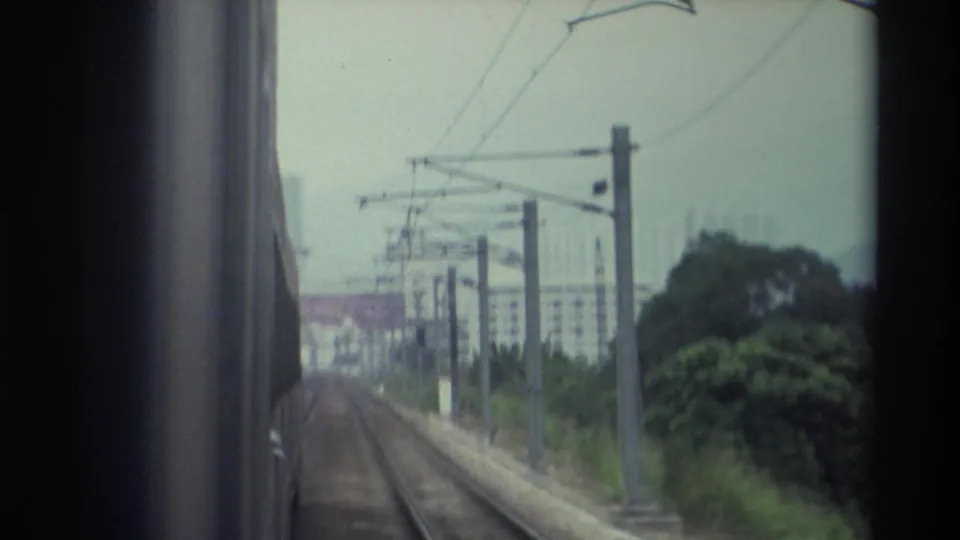 Super 8 train POV, apparently Hong Kong 1982 — prominent electrical transmission pylons and catenary infrastructure flank the track, hazy residential towers beyond.