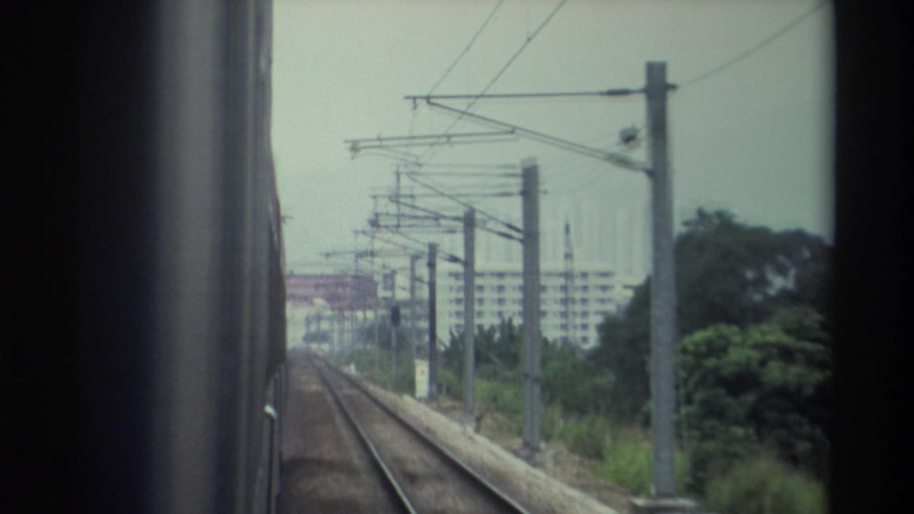 8mm footage from inside a moving train in Hong Kong, circa 1982, looking forward along an open electrified rail line with overhead wires and distant apartment buildings.