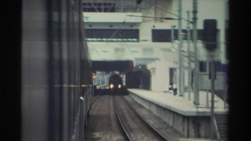 8mm POV from inside a train at a Hong Kong station platform, circa 1982, showing an oncoming train emerging from a tunnel with concrete platform and signage visible.