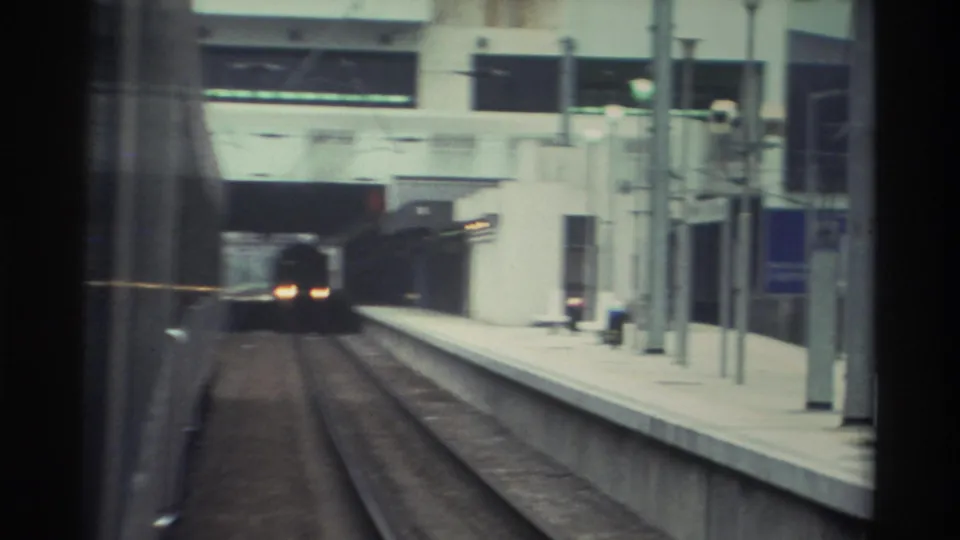 8mm final frame of arriving at a Hong Kong station, circa 1982, with prominent oncoming train headlights and a concrete platform and station canopy framing the view.