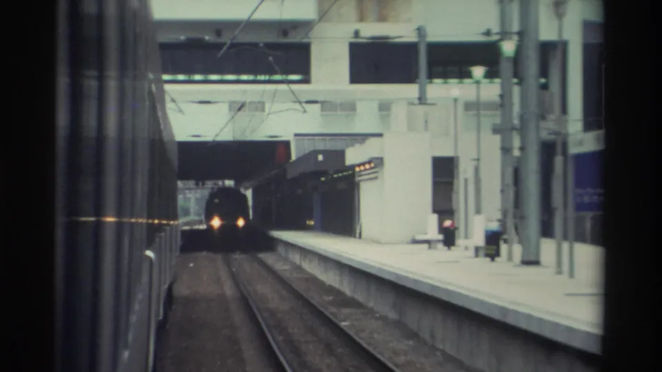 8mm home movie frame, 1982 Hong Kong: POV from a train window looking along a concrete platform as another train approaches with lit headlights.