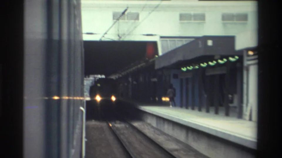 Closing 8mm frame, 1982 Hong Kong transit sequence: oncoming train headlights bright against the tunnel mouth, green platform signals and station canopy at right.
