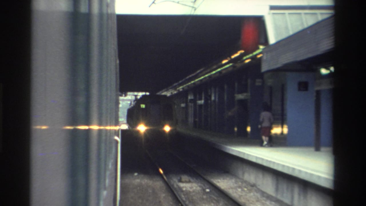 Continuing platform view as train closes in, window frame at left, silhouetted passengers visible under canopy.