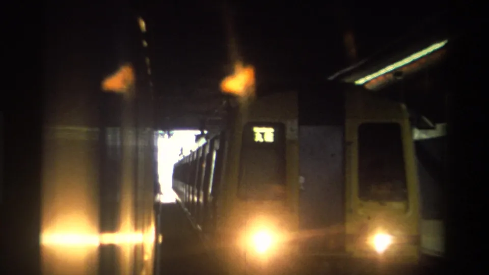 8mm frame from a Hong Kong station tunnel, c. 1982, showing train cars in deep shadow alongside an illuminated green Chinese-character sign with a bright tunnel opening beyond.