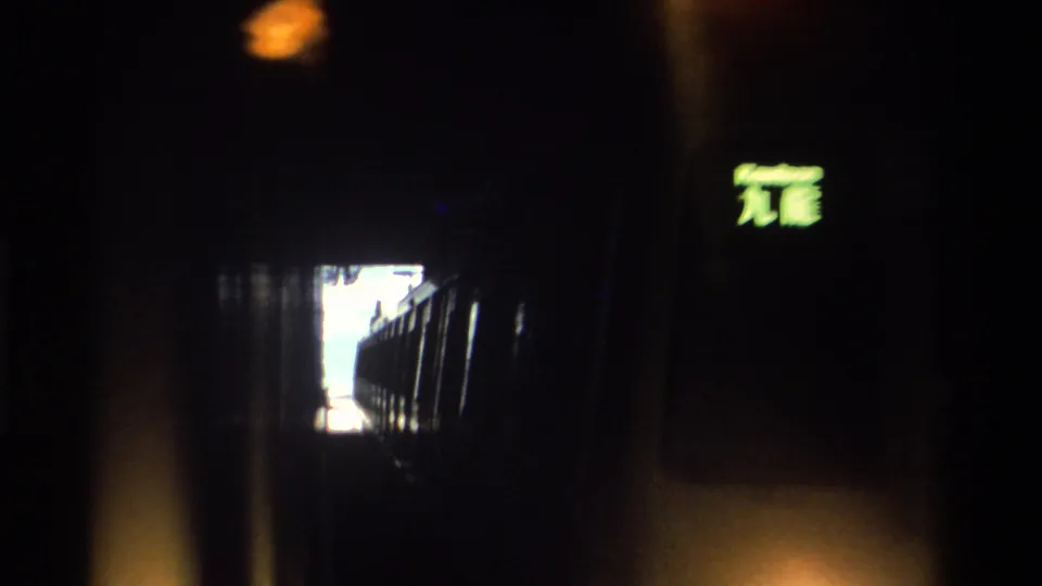 8mm home movie frame from a Hong Kong station tunnel, c. 1982, with a train car in deep shadow, a glowing green Chinese-character sign at upper right, and a lit tunnel opening in center.