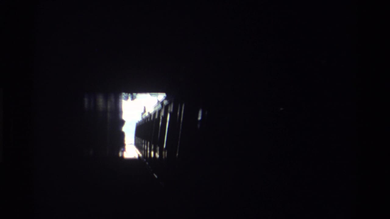 Dark railway tunnel interior with a bright overexposed exit opening at upper-left and dark train structure barely visible in shadow.